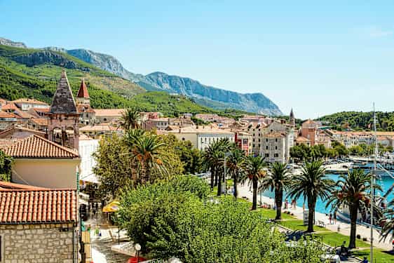 Billedet viser en malerisk middelhavsby med røde tegltage, kirketårne, palmer og grønne bjerge i baggrunden. En lystbådehavn ses også ved vandet. (AI-genereret tekst)