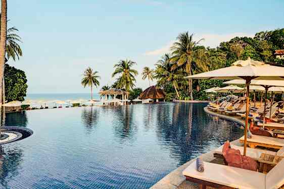 En infinity-pool omgivet af liggestole og parasoller med udsigt over det tropiske landskab og havet. (AI-genereret tekst)