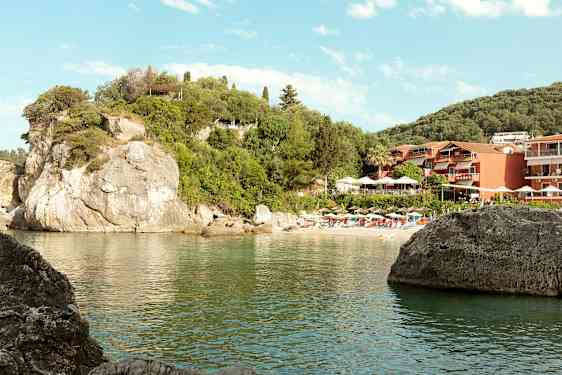Billede af en klippestrand med parasoller og liggestole samt bygninger omgivet af grønt. (AI-genereret tekst)
