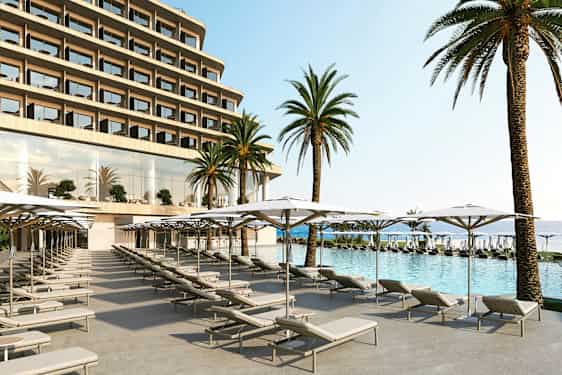 Billedet viser en hotelpool med liggestole og parasoller i forgrunden samt store palmer og hotellets facade i baggrunden. (AI-genereret tekst)