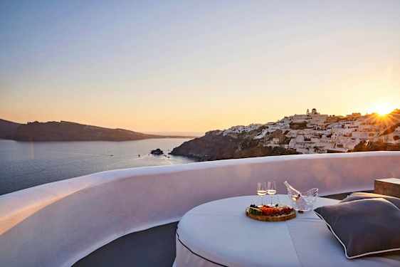 Udsigt over en solnedgang ved havet med hvide bygninger på en klippe og en terrasse med bord, mad og drikke i forgrunden. (AI-genereret tekst)