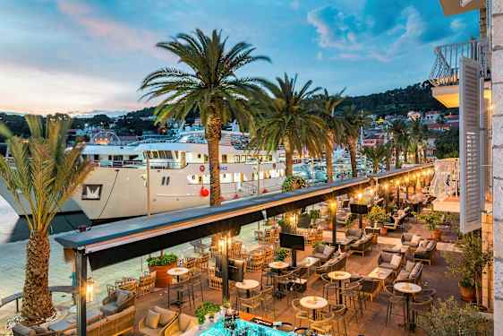 En udsigt over en udendørsrestaurant ved en havn med palmer og store både. (AI-genereret tekst)