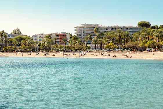 En kyststrækning med klart blåt vand og solbadende mennesker. I baggrunden ses palmer og bygninger. (AI-genereret tekst)