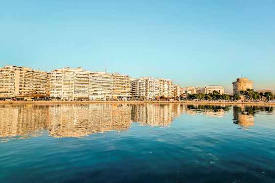 En by ved vandet med bygninger og et rundt tårn reflekteret i det rolige vand. (AI-genereret tekst)