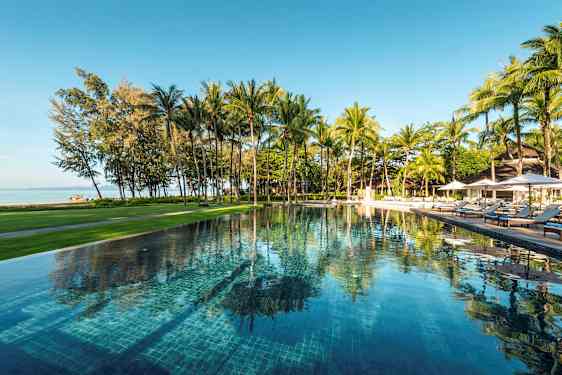 En udendørs swimmingpool omgivet af grønne områder og palmer, med udsigt over stranden og havet. (AI-genereret tekst)