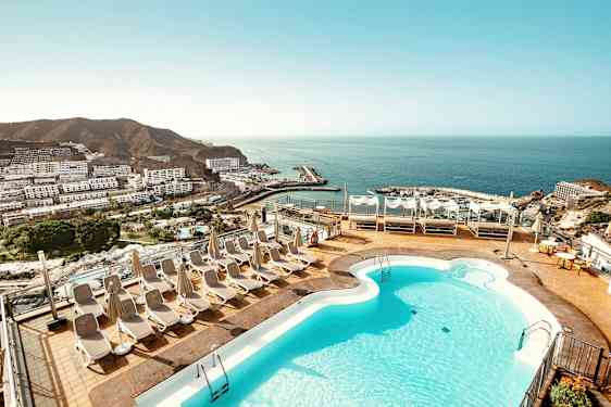 En terrasse med en pool og liggestole, med udsigt over havet og en kystby. (AI-genereret tekst)