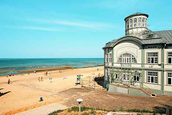 En strand med solbadende mennesker ved en rolig kystlinje, samt en større bygning med klassisk arkitektur ved stranden. (AI-genereret tekst)