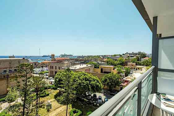 Udsigt fra en altan over en by med historiske bygninger, træer og en havn med både. (AI-genereret tekst)