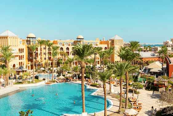 Et billede af et hotelområde med en stor pool, omgivet af palmer og bygninger i varme farver. (AI-genereret tekst)