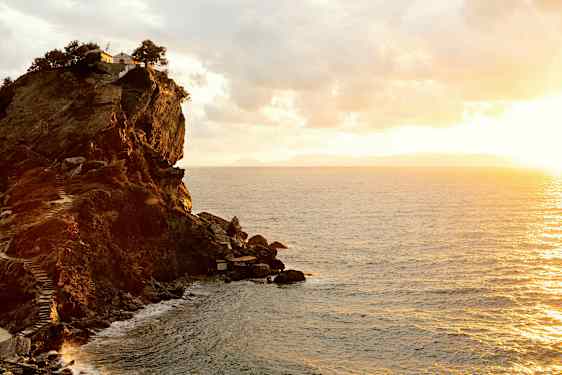En klippe ved kysten med en bygning og træer på toppen. Havet ses under en solnedgang. (AI-genereret tekst)
