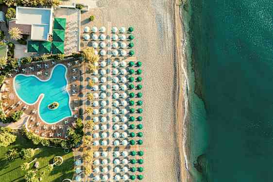 Luftfoto af en hotelpool med omkringliggende liggestole, ved siden af en strand med parasoller og hav. (AI-genereret tekst)