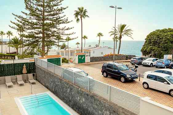 Et billede af et udendørsmiljø med en pool i forgrunden, parkerede biler, træer, bygninger og udsigt over havet i baggrunden. (AI-genereret tekst)