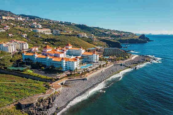 Et hotel med røde tegltage ved havet, omgivet af grønne bakker. (AI-genereret tekst)
