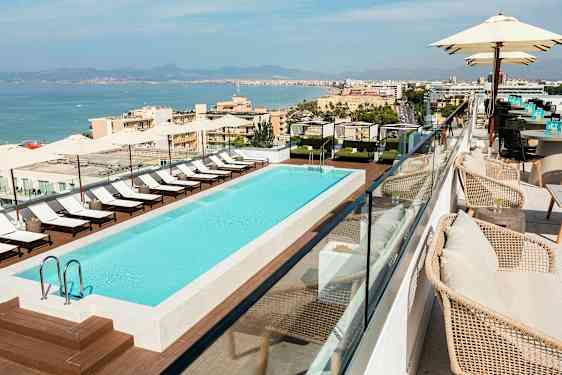 Taglejlighed med en udendørs pool omgivet af liggestole og skygge, med udsigt over en kystby. (AI-genereret tekst)