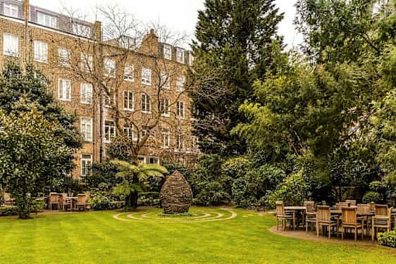 En grøn have med borde og stole samt et stenmonument i midten, omgivet af bygninger. (AI-genereret tekst)