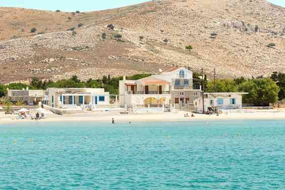 Billede af en klarblå havbugt med en sandstrand samt huse og bakker i baggrunden. (AI-genereret tekst)