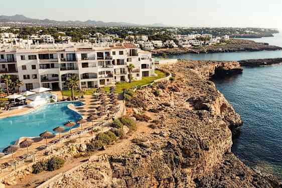 Billedet viser en hotelbygning ved en klippekyst med poolområde og liggestole tæt på havet. (AI-genereret tekst)