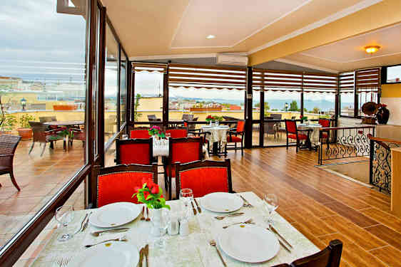 En restaurant med borde dækket til måltid, røde stole og udsigt over en terrasse samt havet. (AI-genereret tekst)