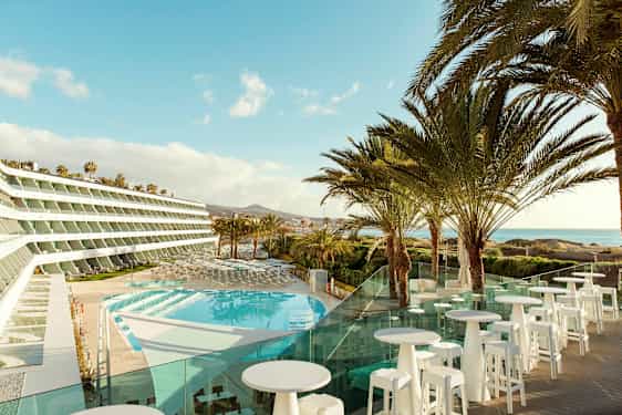 Billede af et hotel med udsigt over en pool og palmer, med havet i baggrunden. (AI-genereret tekst)