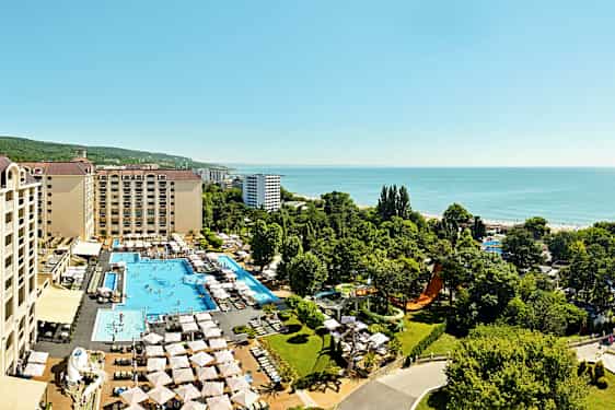 Luftfoto af hotelkompleks med pools, liggestole og grønne områder, beliggende tæt på havet. (AI-genereret tekst)