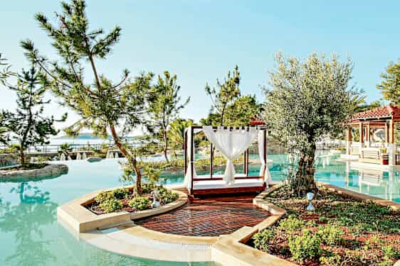 En pool omgivet af træer og planter med en udendørs dagseng og en gangsti i forgrunden. (AI-genereret tekst)