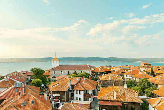 Den gamle bys tage med huse med røde teglstenstage og en kirke i baggrunden nær havet ved kysten. (AI-genereret tekst)