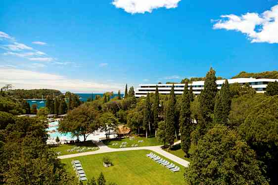 Hotelbygning omgivet af fyrretræer og grønne områder med udsigt over havet. (AI-genereret tekst)