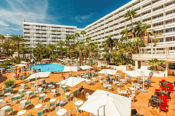 Hotel med stor udendørsterrasse, poolområde og palmer. (AI-genereret tekst)