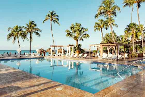 En pool omgivet af solsenge, palmer og en pergola, beliggende tæt på havet med en klar blå himmel. (AI-genereret tekst)