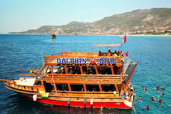 En træbåd med teksten "Dolphin Dive" ligger ved klart blåt vand, hvor dykkere ses i vandet. I baggrunden er bjerge og en kystlinje. (AI-genereret tekst)