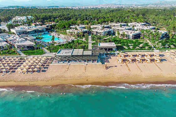 Luftfoto af et strandresort, der viser sandstranden, liggestole, et poolområde og bygninger med omkringliggende grøntområder. (AI-genereret tekst)