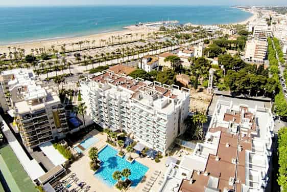 Luftfoto af et hotel med poolområde, omgivet af bygninger og grønne områder. I baggrunden ses en sandstrand og havet. (AI-genereret tekst)