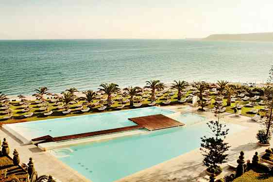 Udsigt over en swimmingpool, liggestole og græsarealer med havet i baggrunden. (AI-genereret tekst)