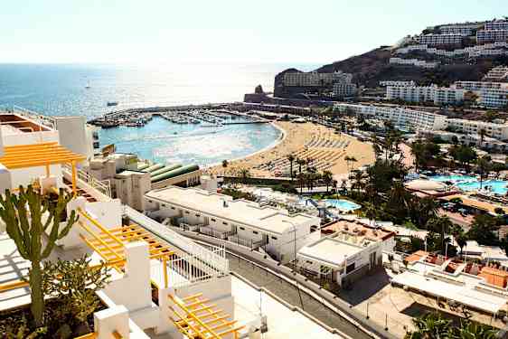 Udsigt over kystby med hvide bygninger, en strand og en lille havn ved havet. (AI-genereret tekst)