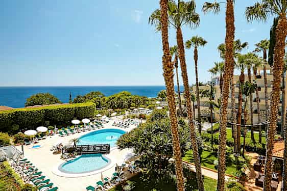 En hotelpool med liggestole og parasoller er omgivet af grønt med havet i baggrunden. (AI-genereret tekst)