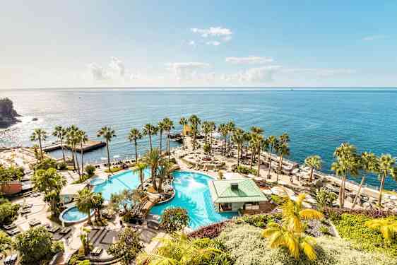 En stor pool med omkringliggende palmer og solsenge, beliggende ved stranden med udsigt over det glitrende hav. (AI-genereret tekst)