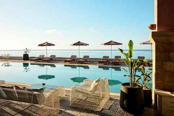 En swimmingpool med liggestole og parasoller ved siden og udsigt over havet. Foran poolen står stole og borde samt store krukkeplanter. (AI-genereret tekst)