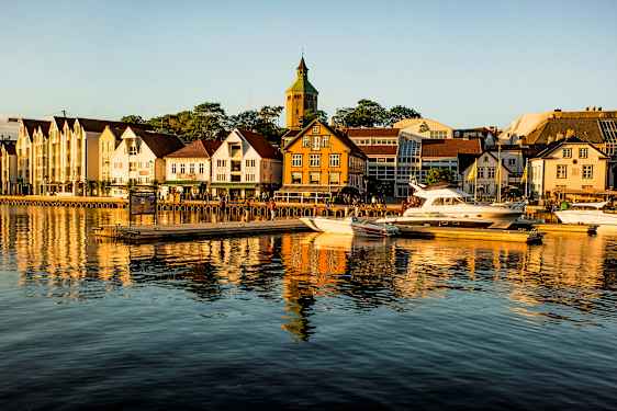 En havn med både liggende ved broen foran bygninger og en kirke i baggrunden. (AI-genereret tekst)