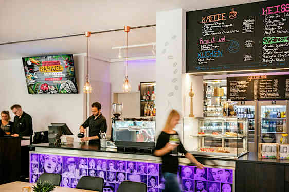 En caféindretning med en disk med en kaffemaskine, bagværk i en glasmontre og en menu på væggen ovenfor. Personale står bag disken. (AI-genereret tekst)