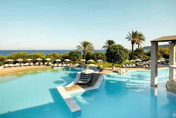 Billede af en stor udendørs pool med liggestole og parasoller omkring, omgivet af grønne områder og palmer med havet i baggrunden. (AI-genereret tekst)