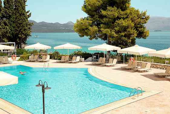 Udendørs swimmingpool med parasoller og liggestole, i baggrunden ses havet og bjergene. (AI-genereret tekst)