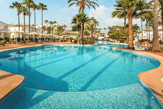 Stor udendørs pool med liggestole og parasoller omgivet af palmer og bygninger. (AI-genereret tekst)