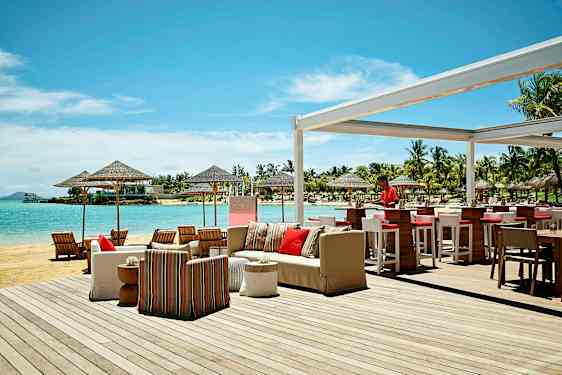 Udendørs serveringsområde ved stranden med bar og siddepladser, samt udsigt over havet og omkringliggende palmer. (AI-genereret tekst)