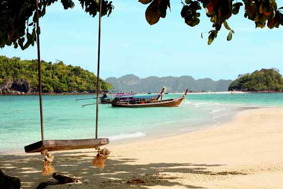 Strand med gynge og en træbåd på vandet foran skovklædte øer. (AI-genereret tekst)