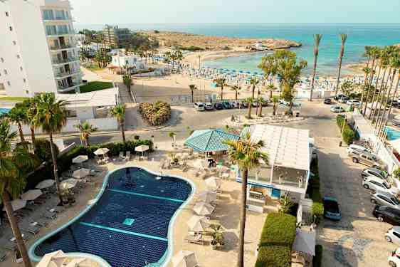 Et overblik over en hotelpool med liggestole, omgivet af palmer, med havet og en solrig strand i baggrunden. (AI-genereret tekst)