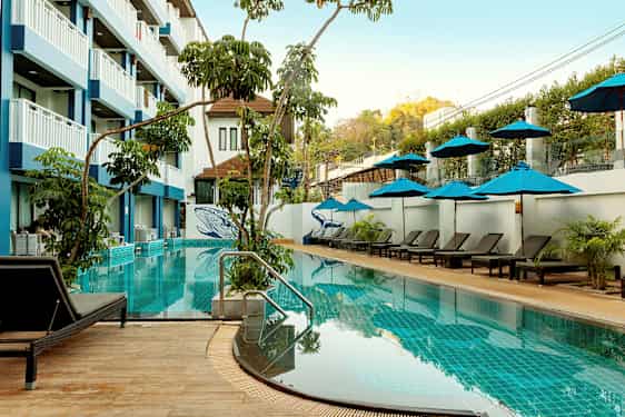 En udendørs swimmingpool med liggestole og parasoller, omgivet af vegetation og en fler-etagers hotelbygning. (AI-genereret tekst)