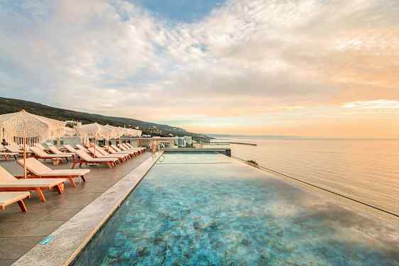 Udsigt over en infinitypool med liggestole og parasoller ved siden af, mod havet ved solnedgang. (AI-genereret tekst)