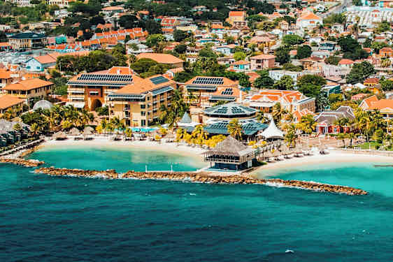 Luftfoto af en kystby med turkis vand, en sandstrand og bygninger med røde tage og solcellepaneler. (AI-genereret tekst)