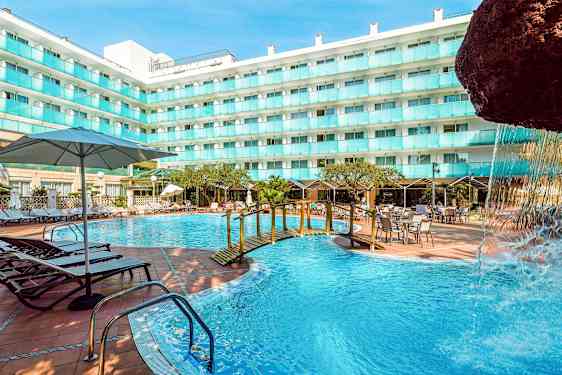 En udendørs swimmingpool ved en hotelbygning med liggestole, borde og parasoller. En lille bro over poolen og en del af en vandfaldsstruktur ses. (AI-genereret tekst)