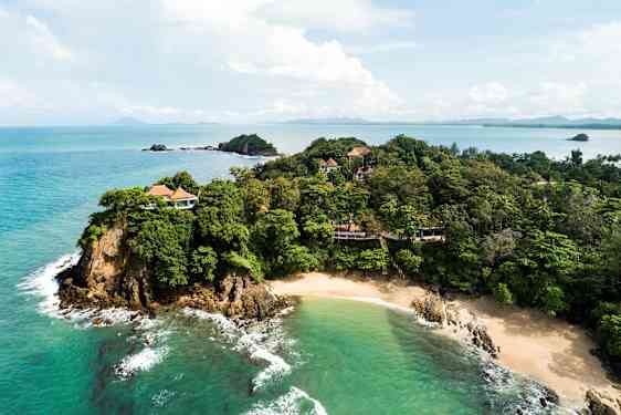 En kystlinje med grønne træer og villaer ses ved siden af en sandstrand og klart turkis hav. (AI-genereret tekst)